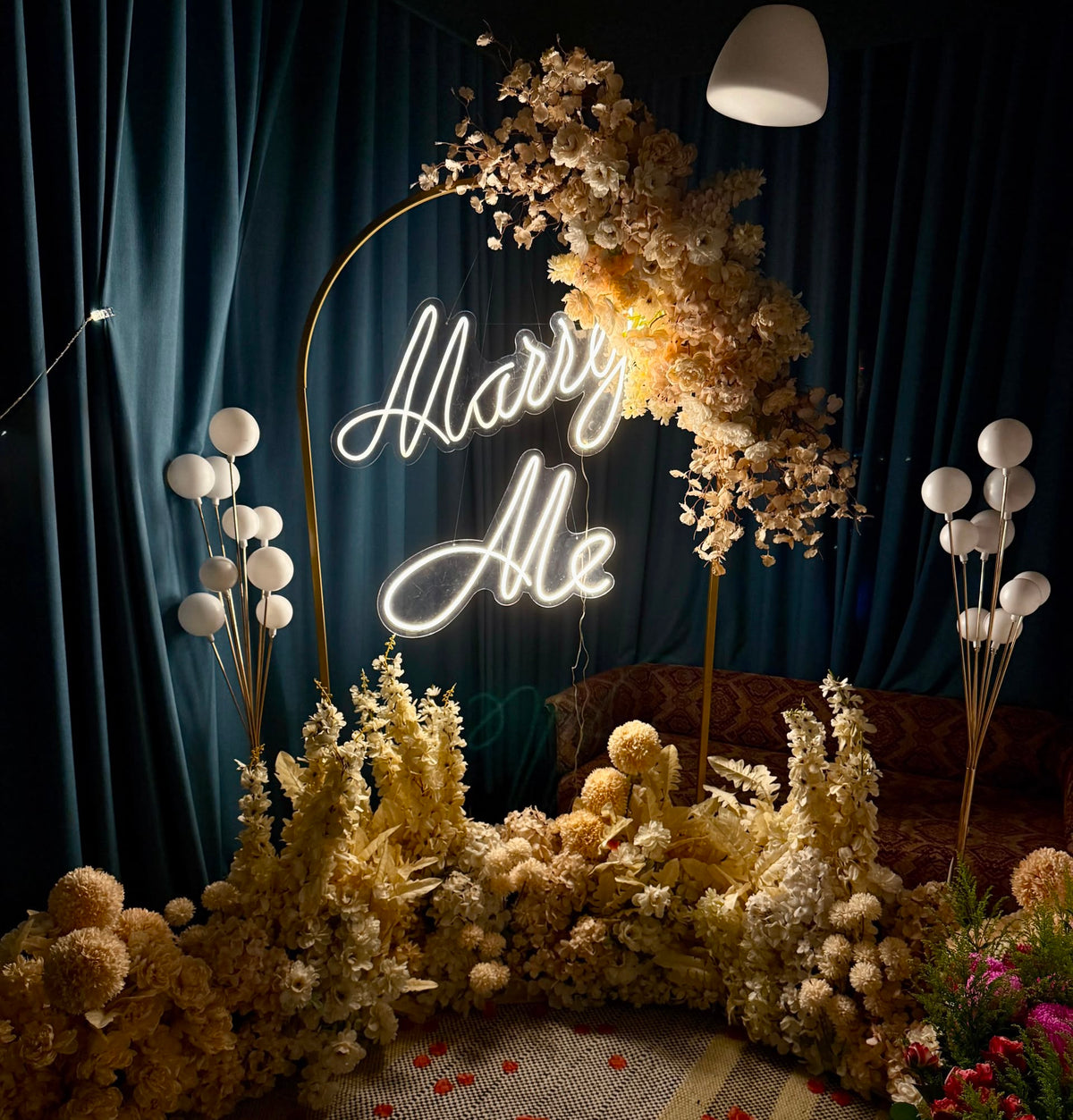 Marry Me proposal backdrop with gold floral arch, neon sign and seating corner
